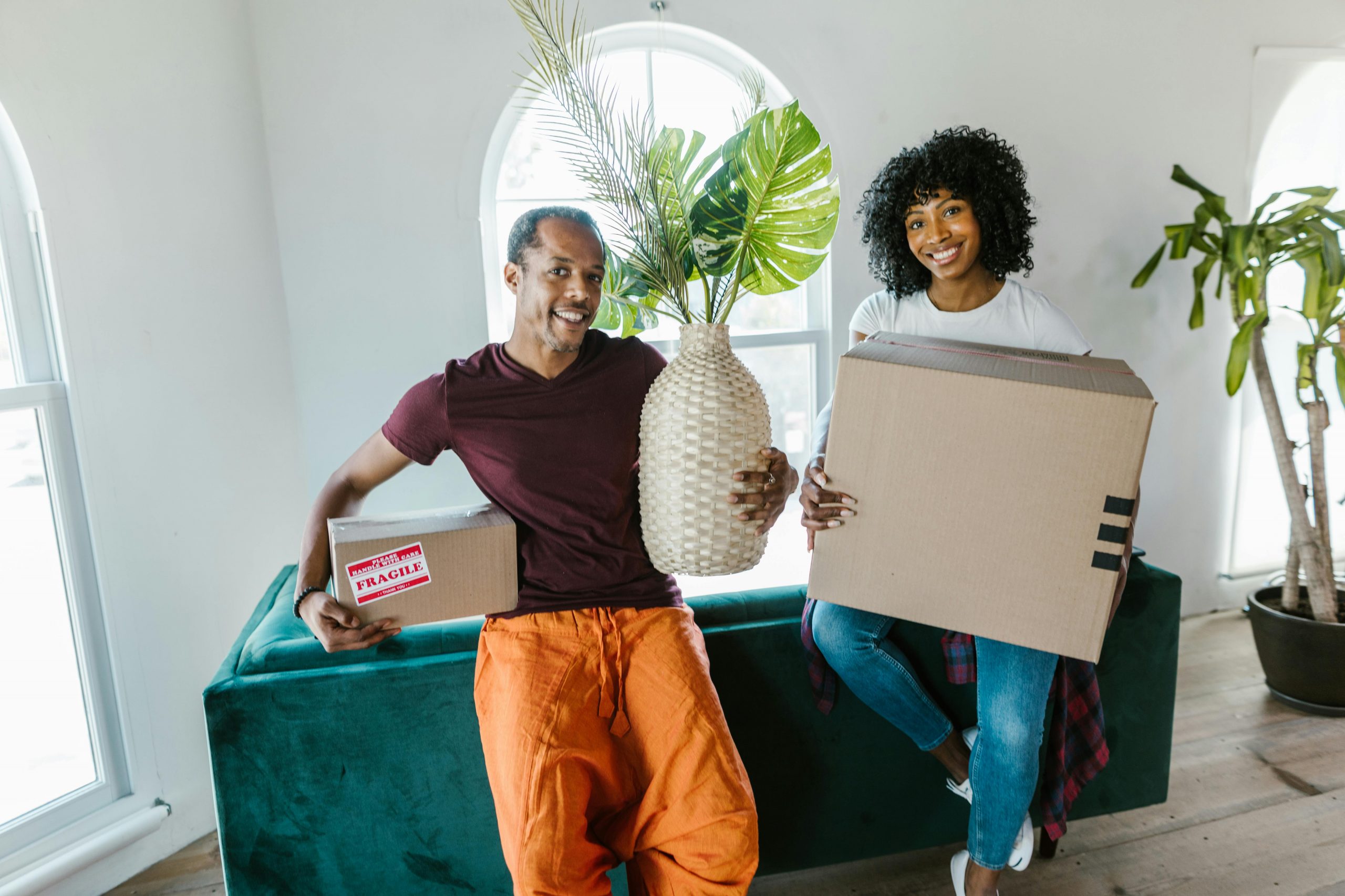 Couple smiling while moving into a new home, carrying boxes and a plant.